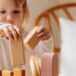 Game Developers - Cute little girl in white casual clothes standing near table and playing with wooden blocks while spending time at home
